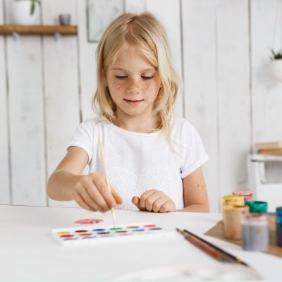 Cute and charming blonde girl wearing white t'shirt deeping brush into paint. Cheerful child with freckles preparing surprise for her parents. Kids and art concept.